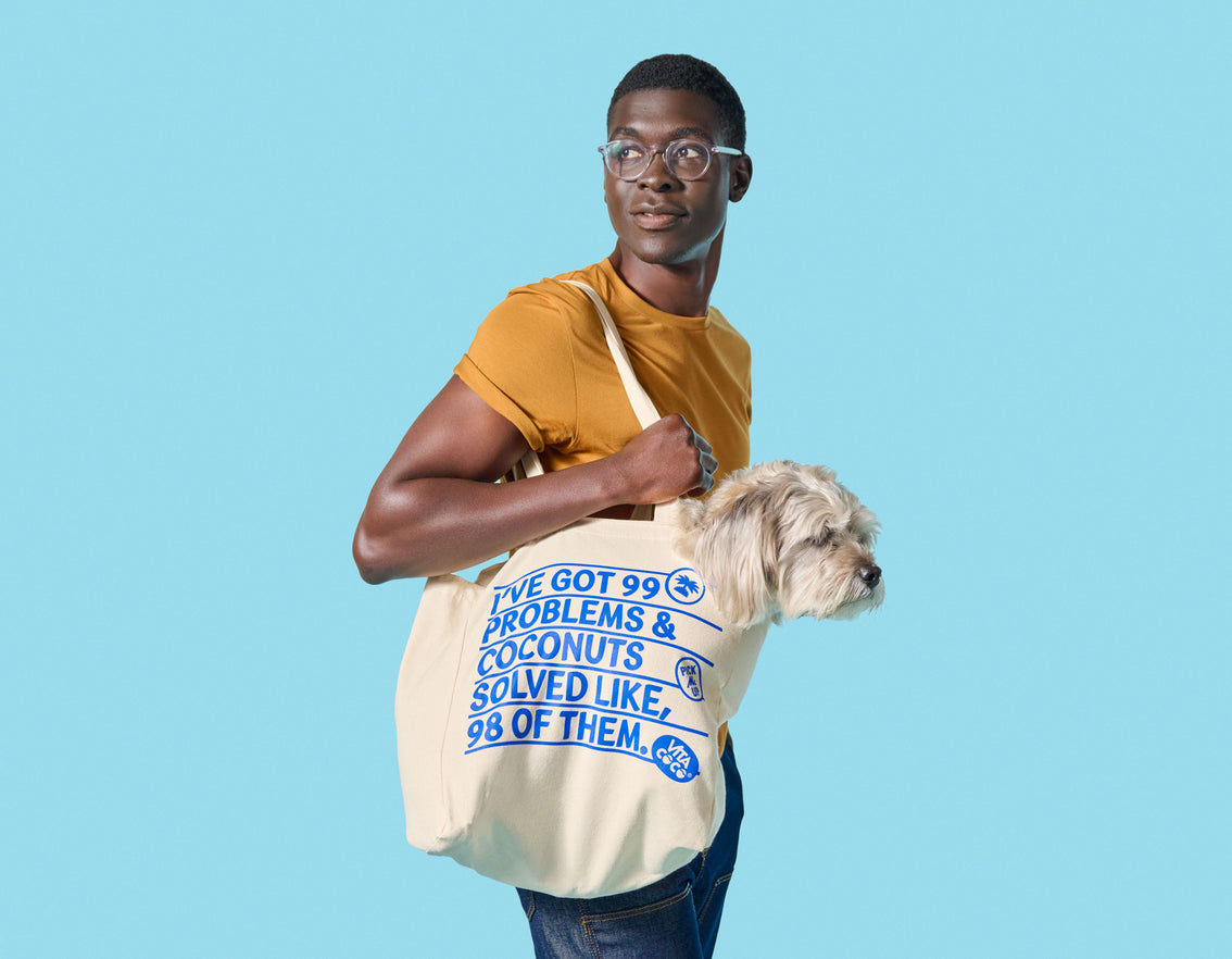 A man carrying a Vita Coco 99 Problems Tote Bag with a dog in it, while shopping at a local store.