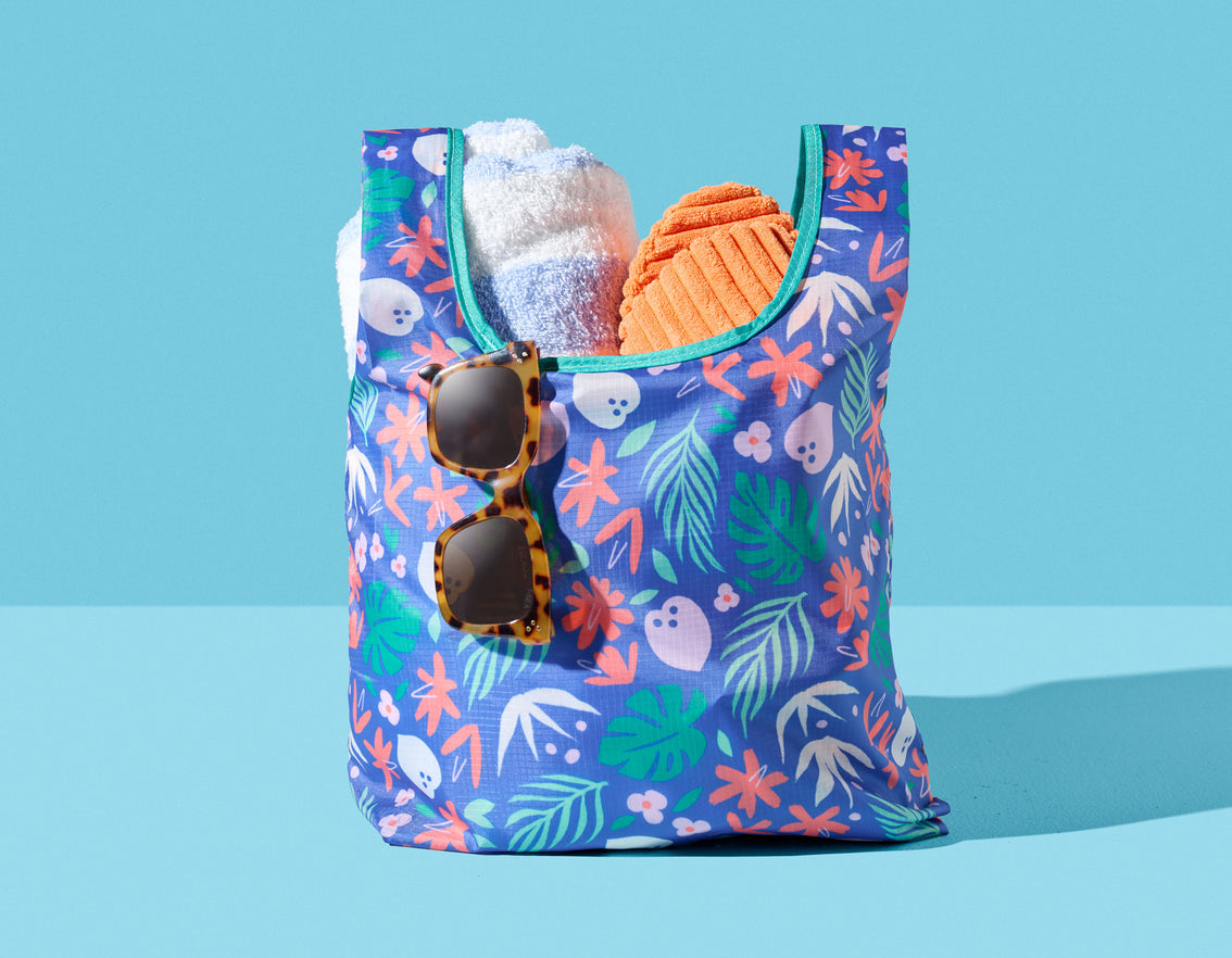 A Mini Patterned Foldable Tote from Vita Coco with a pair of sunglasses and a pair of flip flops that folds easily, perfect for carrying any stuff.